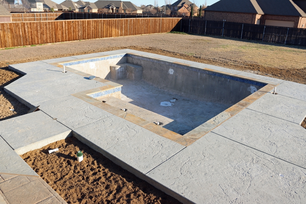 Newly built in-ground pool with a stamped concrete deck awaiting landscaping in a fenced backyard