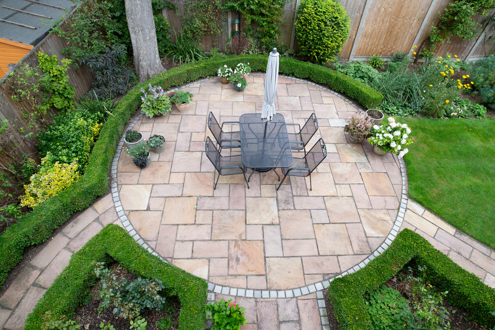 Aerial view of a circular natural stone patio with a dining set, bordered by boxwood hedging and paver pathways