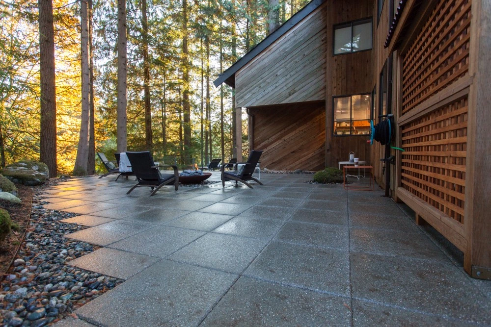 Custom concrete patio with large square pavers beside a modern wood-clad home, featuring outdoor lounge chairs and a fire pit in a forested backyard setting.