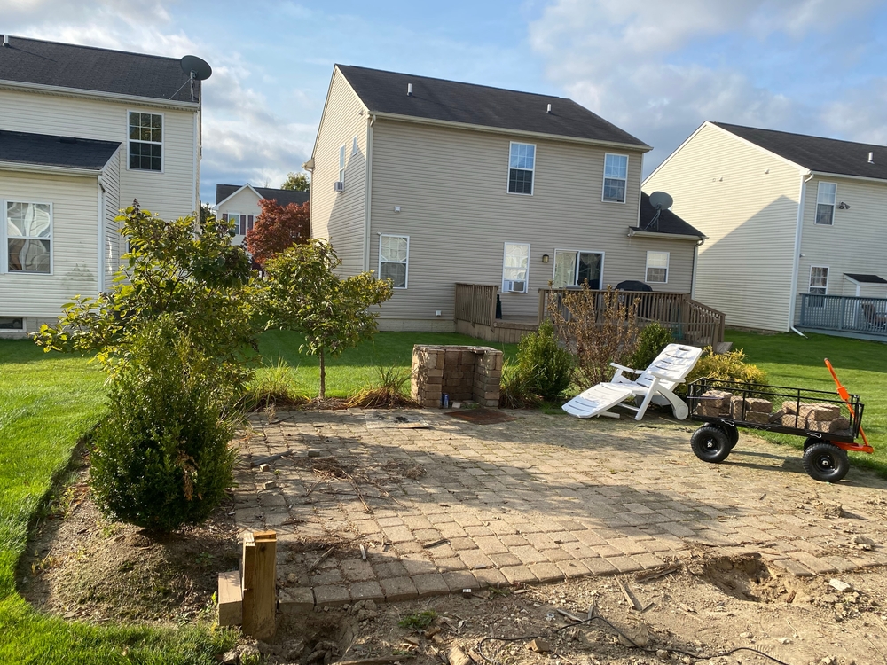 Backyard paver patio installation in progress behind a two-story Langley home with new plantings and a stone fire pit base