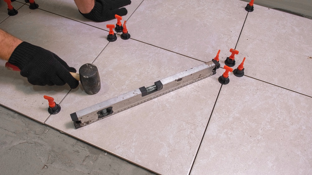 Gloved installer leveling large porcelain floor tiles with a rubber mallet and spirit level